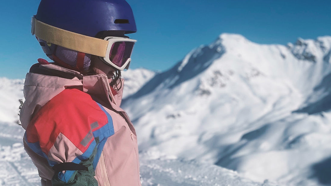 A child is wearing ski goggles, a purple helmet and a pink jacket on a family ski trip.