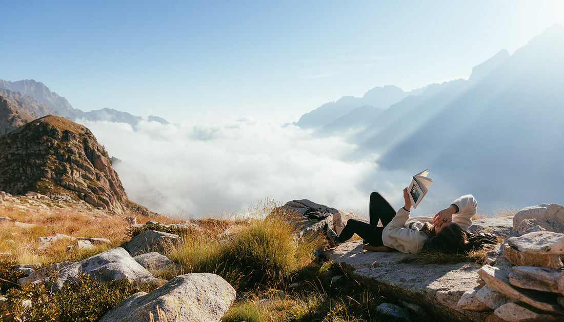 A woman reads a book while relaxing on a rock, the background features mountain tops and a sea of clouds.