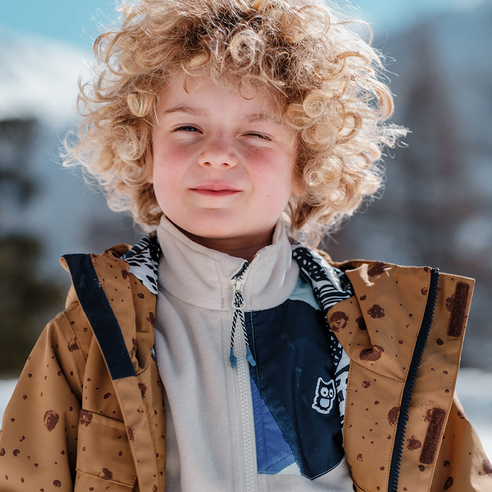 A 5 year old boy with curly blond hair is wearing a brown ski jacket and a cream fleece midlayer.