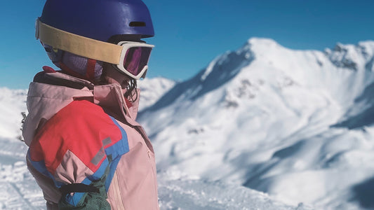 A child is wearing ski goggles, a purple helmet and a pink jacket on a family ski trip. 