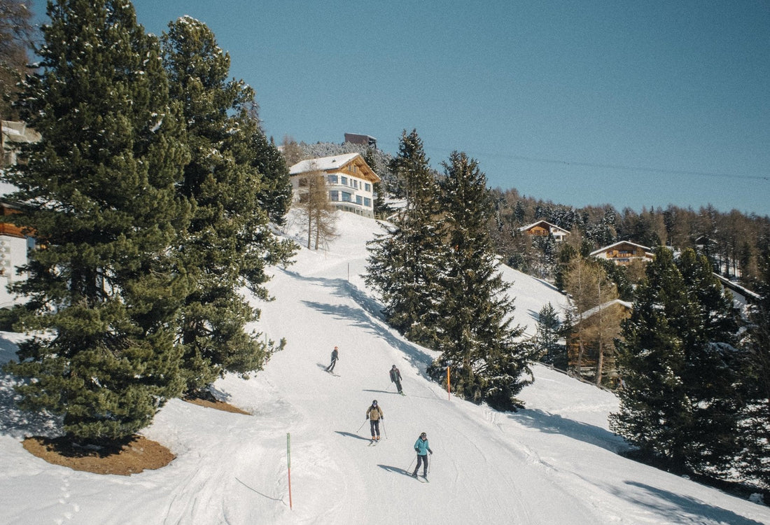 A family of skiers skiing down a narrow but gentle piste between trees and chalets