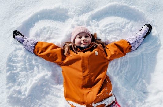 A seven year old girl with a brown jacket and purple ski gloves makes an angel shape in the snow