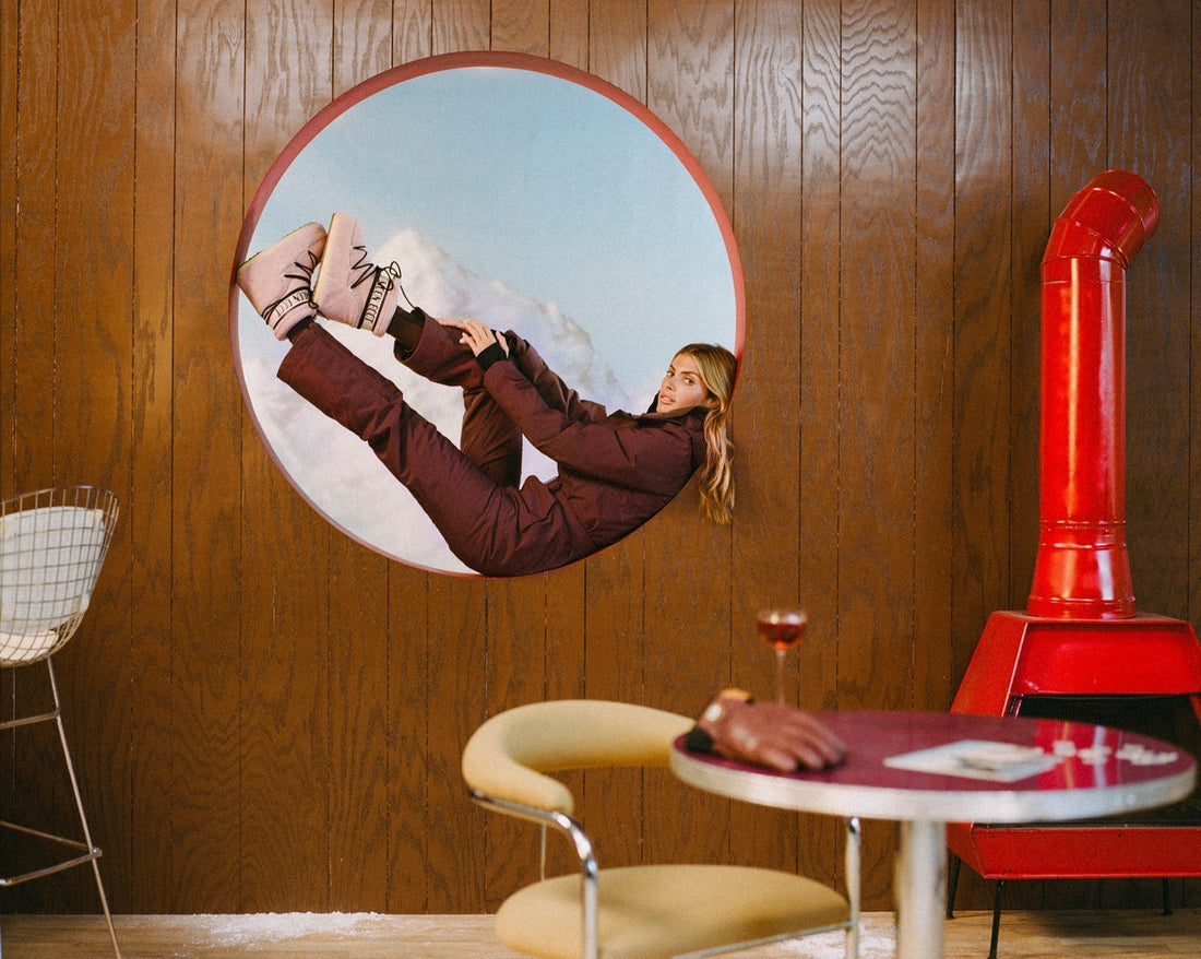 Woman in a dark cherry ski suit puts her legs up while reclining in the window of an alpine cabin