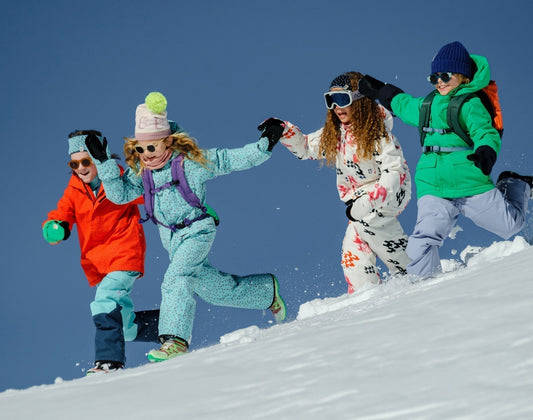 Four kids wearing bright ski suits, colourful hats, glasses and goggles run in the snow holding hands. 
