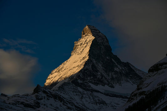 A dramatic shot of the Matterhorn at dusk, the setting sun lights on side of it, with the dark sky closing in. 