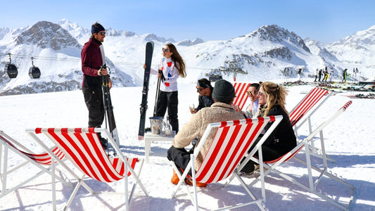 A group of friends take a break from skiing and have a drink on red deck chairs on a snowy terrace. 